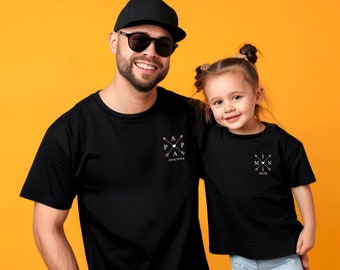 May include: A black t-shirt with the text "Papa" and arrows, and a black t-shirt with the text "Mini" and arrows. The shirts are worn by a man and a girl.