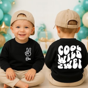 Könnte beinhalten: Ein schwarzes Sweatshirt mit dem Wort "Elias" und einem weißen Peace-Zeichen. Auf der Rückseite des Sweatshirts stehen die Worte "COOL WILD TWO" in Weiß. Das Kind trägt eine beige Baseballkappe und eine beige Hose.