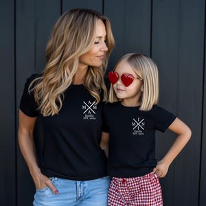 Pu&ograve; includere: Due persone indossano magliette nere con testo bianco. Il testo sulle magliette dice "M & M" con una croce al centro e "Emilia EST. 2019". La persona a sinistra indossa jeans blu e la persona a destra indossa una gonna a quadri rossi e bianchi. La persona a destra indossa occhiali da sole a forma di cuore rossi.