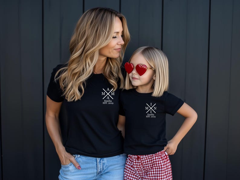 Pu&ograve; includere: Due persone indossano magliette nere con testo bianco. Il testo sulle magliette dice "M & M" con una croce al centro e "Emilia EST. 2019". La persona a sinistra indossa jeans blu e la persona a destra indossa una gonna a quadri rossi e bianchi. La persona a destra indossa occhiali da sole a forma di cuore rossi.