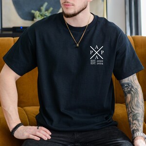 Pu&ograve; includere: Una maglietta nera con un logo bianco che dice "A P P A EST. 2022 EST. 2024". Il logo &egrave; una croce stilizzata con le lettere "A P P A" all'interno.