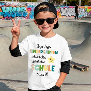 Puede incluir: Una camiseta blanca con el texto "Bye Bye Kindergarten Ich rocke jetzt die SCHULE" en varios colores. La camiseta presenta un gr&aacute;fico de estrella sonriente y el nombre "Elias". La persona lleva una gorra negra y gafas de sol.