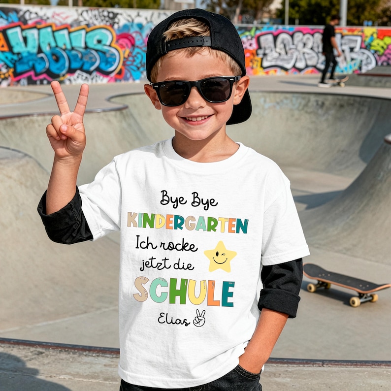 Puede incluir: Una camiseta blanca con el texto "Bye Bye Kindergarten Ich rocke jetzt die SCHULE" en varios colores. La camiseta presenta un gr&aacute;fico de estrella sonriente y el nombre "Elias". La persona lleva una gorra negra y gafas de sol.