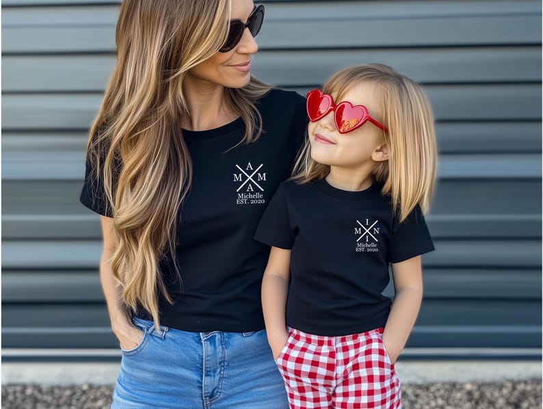 Pu&ograve; includere: Una madre e una figlia indossano magliette nere abbinate con un design grafico bianco. Il grafico presenta una croce con le lettere "MAM" e "MIN" su ogni lato. Il testo "Michelle EST. 2020" &egrave; sotto la croce. La figlia indossa occhiali da sole rossi a forma di cuore.