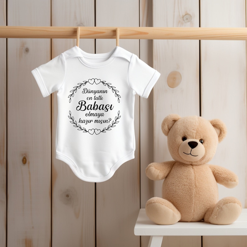 May include: White baby onesie with black text that reads "Dunyannın en tatlı Babası olmaya hazır mısın?"  The onesie is hanging on a wooden hanger. A brown teddy bear is sitting on a white stool.