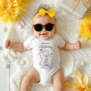 Puede incluir: Un beb&eacute; con un body blanco con un dibujo de oso, gafas de sol y una diadema amarilla con lazo. El body tiene el texto "UNSER ERSTER Muttertag" y "Mama & Lilly". El beb&eacute; est&aacute; acostado sobre una manta blanca con flores amarillas y una caja de regalo.