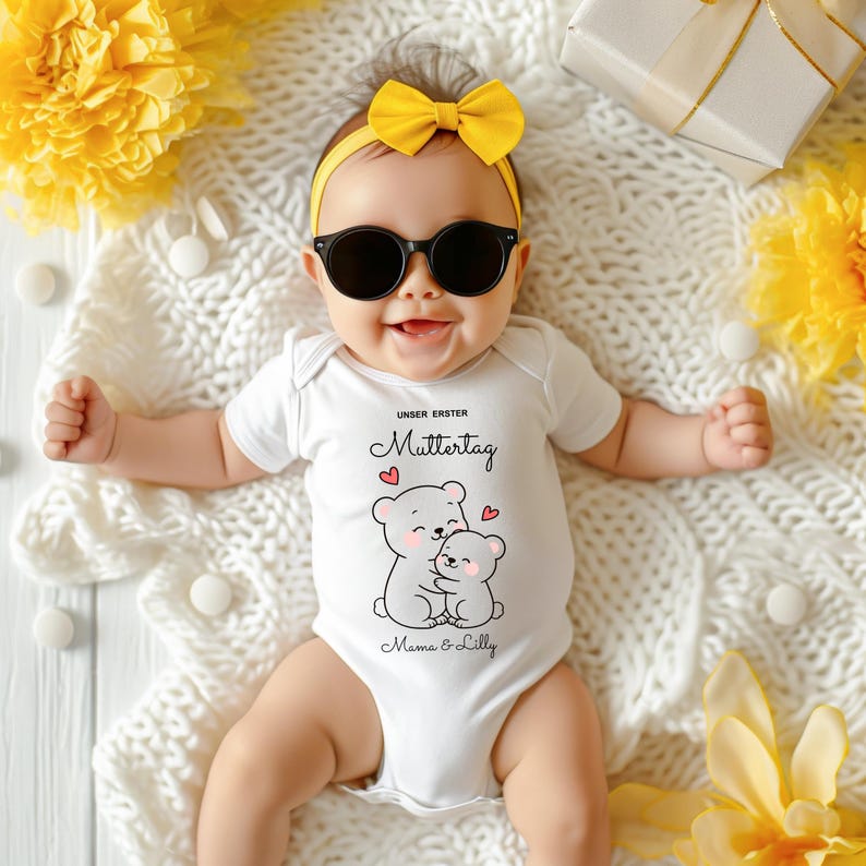 Puede incluir: Un beb&eacute; con un body blanco con un dibujo de oso, gafas de sol y una diadema amarilla con lazo. El body tiene el texto "UNSER ERSTER Muttertag" y "Mama & Lilly". El beb&eacute; est&aacute; acostado sobre una manta blanca con flores amarillas y una caja de regalo.