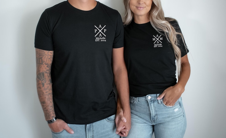 Pu&ograve; includere: Due magliette nere con testo bianco. La maglietta a sinistra dice "Papa" con una croce e il testo "Michelle Est. 2004". La maglietta a destra dice "Mama" con una croce e il testo "Michelle Est. 2004".