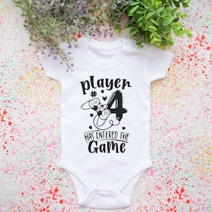 May include: White baby grow with black text that reads "Player #4 has entered the game". The number 4 is styled to look like a video game controller.