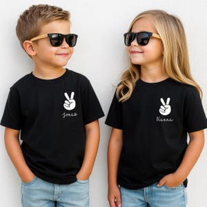 May include: Two black t-shirts featuring a white graphic of a hand making a peace sign on the left chest. Below the peace sign, the left shirt has the name Jonas printed in white script, and the right shirt has the name Hanna printed in white.