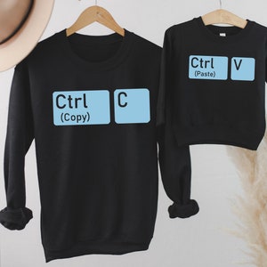 May include: Two black sweatshirts with blue computer keys printed on the front. The larger sweatshirt has the keys "Ctrl" and "C" with the text "(Copy)" below. The smaller sweatshirt has the keys "Ctrl" and "V" with the text "(Paste)" below.