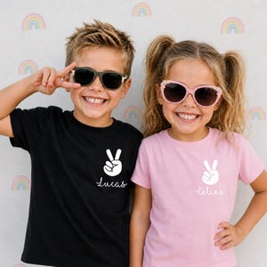 May include: A black t-shirt and a light pink t-shirt, each featuring a white graphic of a hand making a peace sign above the name Lucas or Celine in white script. The black shirt displays the name Lucas, while the pink shirt displays the name Celine.