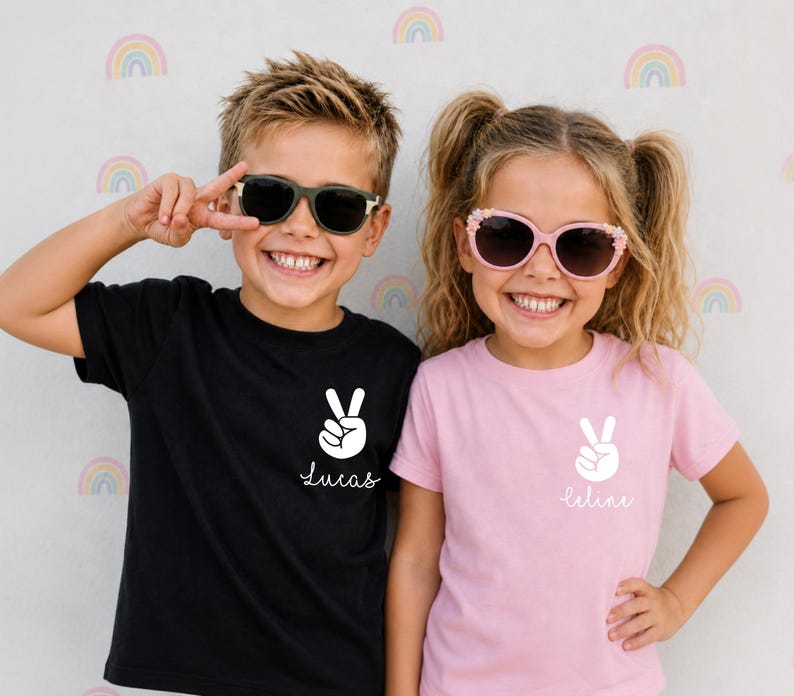 May include: A black t-shirt and a light pink t-shirt, each featuring a white graphic of a hand making a peace sign above the name Lucas or Celine in white script. The black shirt displays the name Lucas, while the pink shirt displays the name Celine.