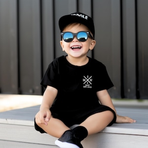 Pu&ograve; includere: Un bambino piccolo che indossa una maglietta nera con una grafica bianca che dice "I M N I Emil EST. 2023". Il bambino indossa anche un berretto da baseball nero con la parola "DOLE" e occhiali da sole neri.