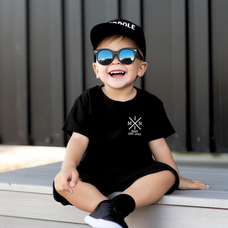 Pu&ograve; includere: Un bambino piccolo che indossa una maglietta nera con una grafica bianca che dice "I M N I Emil EST. 2023". Il bambino indossa anche un berretto da baseball nero con la parola "DOLE" e occhiali da sole neri.