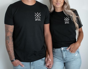 May include: Two black t-shirts with white text. The shirt on the left says "Papa" with a cross and the text "Michelle Est. 2004". The shirt on the right says "Mama" with a cross and the text "Michelle Est. 2004".