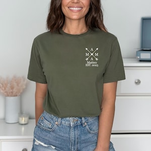 Op de afbeelding: Een groen T-shirt met een wit grafisch ontwerp met de tekst "A Mom A Matteo Est. 2023". De grafiek bevat een hart en pijlen.