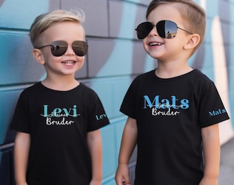 May include: Two young children wearing black t-shirts and sunglasses pose in front of a blue and gray wall. The shirts have the names "Levi" and "Mats" printed in blue and white script, with the words "Bruder" below.