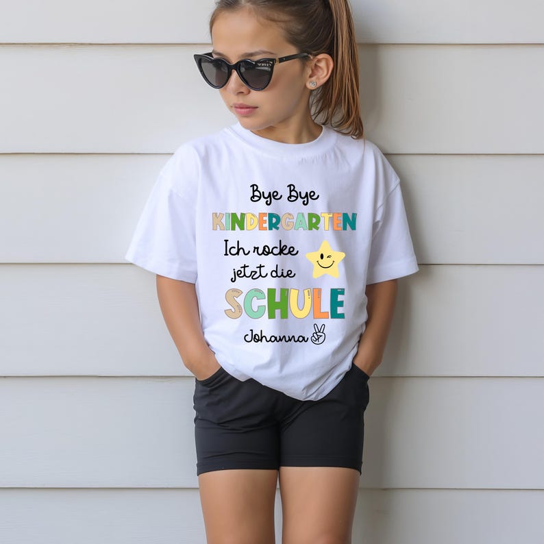 Puede incluir: Camiseta blanca con el texto "Bye Bye Kindergarten Ich rocke jetzt die SCHULE Johanna" en varios colores. La camiseta presenta una estrella amarilla con una cara sonriente. La persona lleva pantalones cortos negros y gafas de sol.