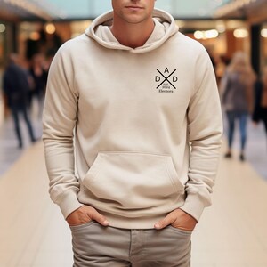 May include: A beige hoodie with a front pocket and a hood. The hoodie has a black graphic on the upper left chest that reads "DAD" with "EST. 2024 Eleonora" underneath. The person is wearing gray pants.