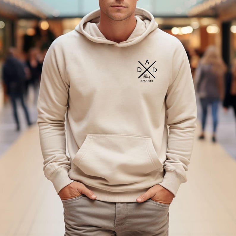 May include: A beige hoodie with a front pocket and a hood. The hoodie has a black graphic on the upper left chest that reads "DAD" with "EST. 2024 Eleonora" underneath. The person is wearing gray pants.