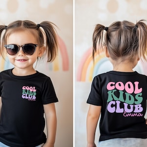 Könnte beinhalten: Schwarzes T-Shirt mit dem Text "COOL KIDS CLUB" in Rosa, Grün und Lila, darunter der Name "Emma". Das T-Shirt wird von einem jungen Kind mit Sonnenbrille getragen. Im Hintergrund ist ein Regenbogen zu sehen.