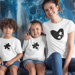 May include: A family wearing white t-shirts with black puzzle piece designs. The adult is wearing a shirt with a heart shape and two puzzle pieces inside. The children are wearing shirts with a single puzzle piece. The family is sitting on a blue couch.