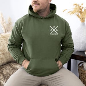 May include: Olive green hoodie with a front pocket and a hood. The chest features a white graphic with the letters "DAD" and the date "20/02/24" with names below. The hoodie is a casual garment, suitable for everyday wear.
