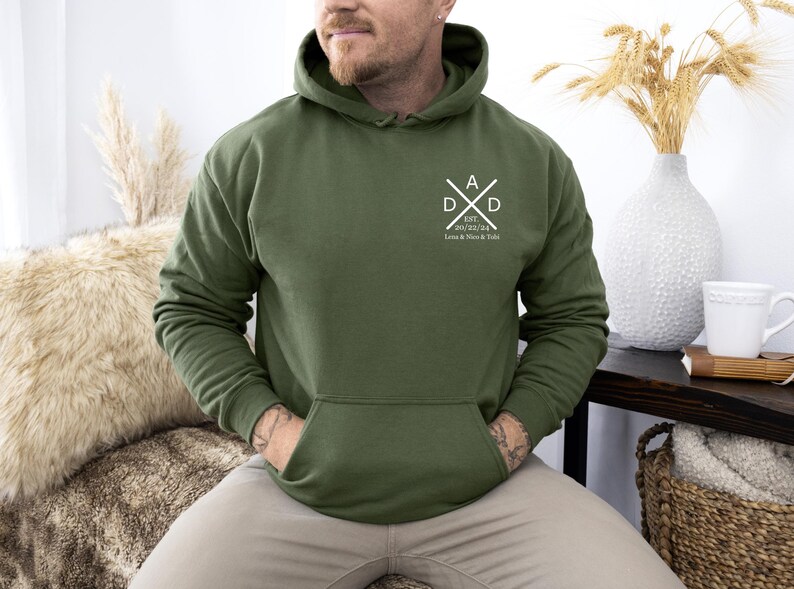May include: Olive green hoodie with a front pocket and a hood. The chest features a white graphic with the letters "DAD" and the date "20/02/24" with names below. The hoodie is a casual garment, suitable for everyday wear.