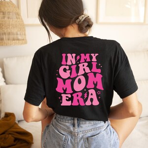 Peut inclure: T-shirt noir avec un texte rose qui dit "In My Girl Mom Era" avec des &eacute;clairs et des &eacute;toiles.