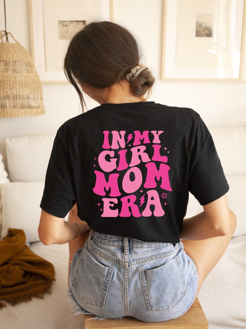 Peut inclure: T-shirt noir avec un texte rose qui dit "In My Girl Mom Era" avec des &eacute;clairs et des &eacute;toiles.