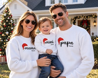 May include: Three white long-sleeve sweatshirts featuring a red and white Santa hat graphic above the black cursive text mama, mini, and papa, with the names Melanie, Jonas, and Thomas printed in black below each respective title.