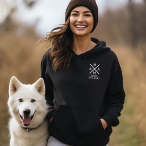 Peut inclure: Un sweat &agrave; capuche noir avec un motif graphique blanc repr&eacute;sentant une croix avec les lettres "A" et "M" de chaque c&ocirc;t&eacute;. Le texte "Louis EST. 2023" est situ&eacute; sous la croix.