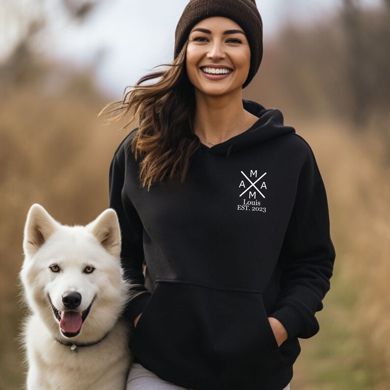 Peut inclure: Un sweat &agrave; capuche noir avec un motif graphique blanc repr&eacute;sentant une croix avec les lettres "A" et "M" de chaque c&ocirc;t&eacute;. Le texte "Louis EST. 2023" est situ&eacute; sous la croix.