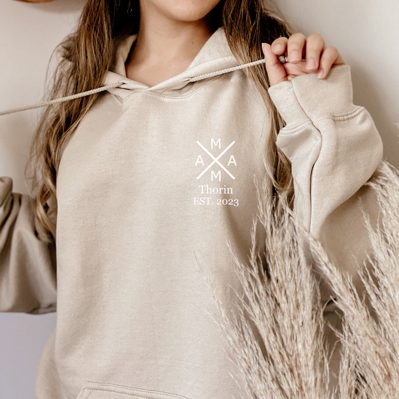 Peut inclure: Un sweat &agrave; capuche beige avec un motif graphique blanc qui dit "MAM" avec une croix au milieu et "Thorin EST. 2023" en dessous.
