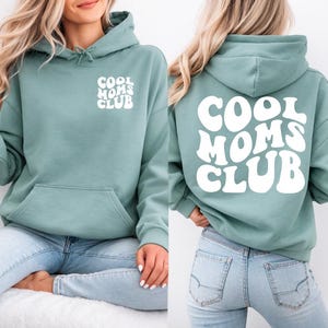 May include: A sage green hooded sweatshirt featuring a front kangaroo pocket and a drawstring hood. The text COOL MOMS CLUB is printed in white on the left chest and in a larger, bold, wavy font across the entire back of the garment.