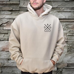 May include: A beige hoodie with a front pocket and a hood. The hoodie features a black graphic on the chest that says "DAD" with crossed lines, the year "20/24", and the names "Nelia & Nico".