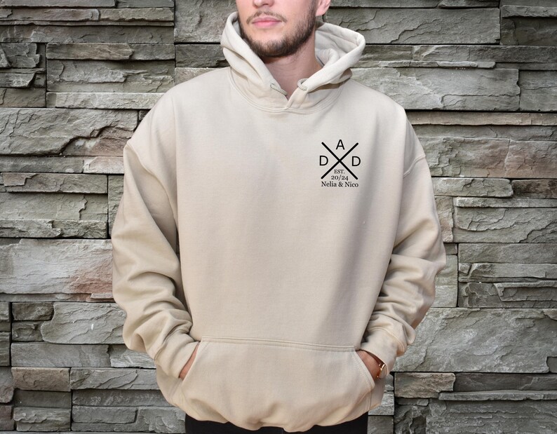 May include: A beige hoodie with a front pocket and a hood. The hoodie features a black graphic on the chest that says "DAD" with crossed lines, the year "20/24", and the names "Nelia & Nico".