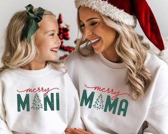 Könnte beinhalten: Zwei Personen tragen passende weiße Sweatshirts mit den Wörtern "merry MAMA" und "merry MINI" in Rot und Grün. Die Person rechts trägt einen roten Weihnachtsmannhut. Die Person links hat eine grüne Schleife im Haar. Weihnachts-Themenbekleidung.