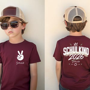 May include: A maroon t-shirt featuring a white peace sign graphic and the name Jonas on the front. The back displays the text SCHULKIND 2026, the name Jonas, a crown icon, and a smiley face. The shirt is paired with a tan and brown baseball cap.