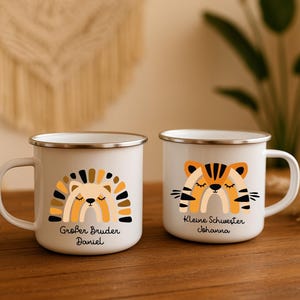 May include: Two white enamel mugs with silver rims. The left mug features a stylized lion graphic with a rainbow mane and the text Gro&szlig;er Bruder Daniel. The right mug features a stylized tiger graphic with a rainbow body and the text Kleine Schwester Johanna.