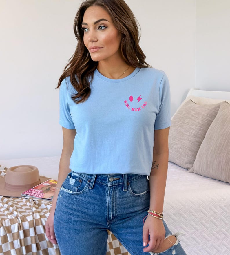 Peut inclure: Un t-shirt bleu clair avec un graphique rose et blanc d'un visage souriant avec un &eacute;clair. Le texte "Girl Mom" est &eacute;crit autour du visage souriant.