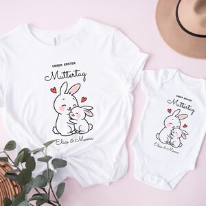 Puede incluir: Camisetas blancas y un body de beb&eacute; con un gr&aacute;fico de conejito abrazando a otro conejito, corazones rojos y el texto "UNSER ERSTER MUTTERTAG" y "Elias & Mama". Un sombrero marr&oacute;n est&aacute; en el fondo.