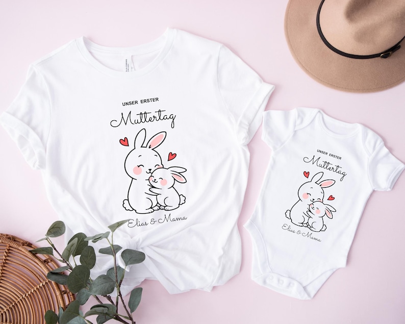 Puede incluir: Camisetas blancas y un body de beb&eacute; con un gr&aacute;fico de conejito abrazando a otro conejito, corazones rojos y el texto "UNSER ERSTER MUTTERTAG" y "Elias & Mama". Un sombrero marr&oacute;n est&aacute; en el fondo.