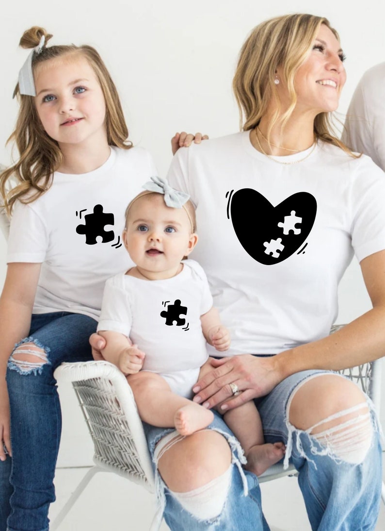 May include: A family of three wearing white t-shirts with black puzzle piece designs. The adult shirt has a black heart with two white puzzle pieces inside. The shirts are a matching set.
