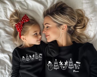 May include: A mother and child are lying on a white surface, wearing matching black sweatshirts. The sweatshirts feature a white Christmas-themed design with a snowman, trees, a reindeer, and a gingerbread man. The child's sweatshirt says "Mama". The child wears a red headband with white polka dots.