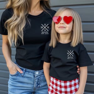 Pu&ograve; includere: Una maglietta nera con un design grafico bianco che dice "A M I N Michelle EST. 2020". Un bambino indossa una maglietta nera abbinata con un design grafico bianco che dice "M I N Michelle EST. 2020". Il bambino indossa pantaloncini a quadri rossi e bianchi e occhiali da sole a forma di cuore.