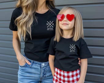 May include: A black t-shirt with a white graphic design that says "A M I N Michelle EST. 2020". A child is wearing a matching black t-shirt with a white graphic design that says "M I N Michelle EST. 2020". The child is wearing red and white checked shorts and heart-shaped sunglasses.