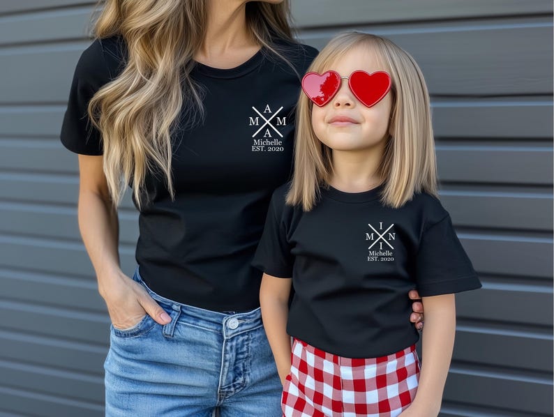 Pu&ograve; includere: Una maglietta nera con un design grafico bianco che dice "A M I N Michelle EST. 2020". Un bambino indossa una maglietta nera abbinata con un design grafico bianco che dice "M I N Michelle EST. 2020". Il bambino indossa pantaloncini a quadri rossi e bianchi e occhiali da sole a forma di cuore.
