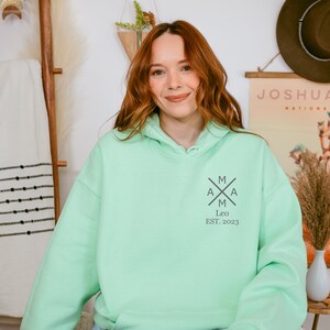 Peut inclure: Un sweat &agrave; capuche vert clair avec un logo noir et blanc qui dit "MAMA" avec une croix au milieu et "Leo EST. 2023" en dessous.
